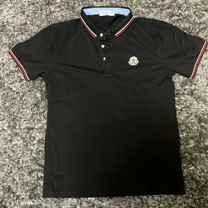 Svart Moncler piké - Svart pikéskjorta från Moncler med klassisk krage och knappar framtill. Röd och vit randig detalj på krage och ärmslut, samt Moncler-logga på bröstet. Skjortan är kortärmad och har en stilren, sportig look.