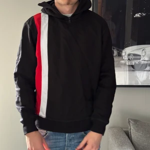 Svart hoodie med röd och vit stripe - Svart hoodie med huva och half zip, prydd med en bred röd och vit stripe längs vänster sida fram och bak. På vänster ärm finns ett broderat märke med siffrorna 1312 och ett kors i rött och vitt. Tröjan har lång ärm och normal passform, perfekt för dig som gillar ultras stilen