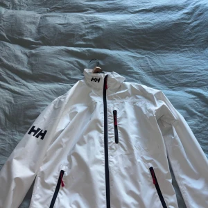 Vit vindjacka från Helly Hansen - Snygg vit vindjacka från Helly Hansen med svarta detaljer och röda dragkedjor. Jackan har hög krage, tre fickor med dragkedja och HH-logga på ärm och krage. Jag har lyckats hålla den vit och hittar inga synliga fläckar. 