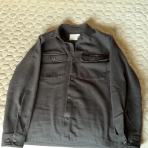 Svart overshirt från Jack & Jones - Stilren svart overshirt från Jack & Jones med klassisk krage, två bröstfickor med knappar och hel knappslå framtill. Jackan har lång ärm och är tillverkad i ett mjukt material som passar perfekt för lager-på-lager-stil. Inga defekter.