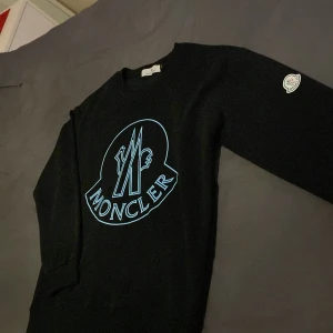 Svart sweatshirt från Moncler - Svart sweatshirt från Moncler med stort blått logotryck på bröstet och klassisk Moncler-patch på ärmen. Tröjan har rund hals, ribbade muddar och är långärmad. Perfekt för dig som gillar stilrena designer med tydlig branding.