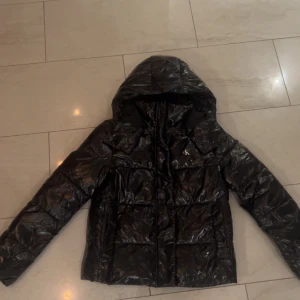 Svart pufferjacka från Calvin Klein - Säljer en svart glansig pufferjacka från Calvin Klein med huva och dragkedja framtill. Jackan har quiltad design och CK-logga på bröstet. Perfekt för kalla dagar och har en cool, oversized vibe. Passar dig som gillar streetstyle och vill hålla dig varm.