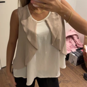 Beige och vit blus  - Snygg blus i beige och vitt med omlottkänsla framtill. Västen har ett flowy, löst fall och är tillverkad i ett tunt, glansigt material som påminner om siden. Rund halsringning och öppen design utan knäppning. Perfekt för att lyfta en enkel outfit.