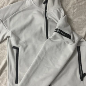 Vit kofta från Sail Racing - Snygg vit kofta från Sail Racing med svarta detaljer och dragkedjor. två sidofickor med dragkedja och logotyp på ärmen. Perfekt för dig som gillar stilren och sportig look. 