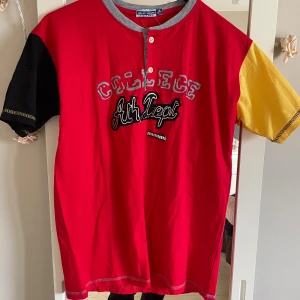 College t-shirt från Relax Mode - Cool t-shirt från Relax Mode med röd kropp, svart och gul ärm samt grå krage. Tryck och broderi på bröstet med texten 'College Ath. Dept'. Kortärmad modell i mjuk bomull, perfekt för en avslappnad stil.