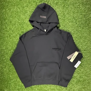 Svart Essentials Fear of God hoodie - Svart hoodie från Essentials Fear of God med stor logga på ryggen och mindre tryck på bröstet. Klassisk känguruficka framtill, huva och ribbade muddar. Perfekt för dig som gillar streetwear och vill ha en clean look.