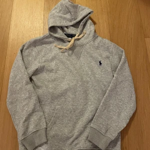 Grå hoodie från Polo Ralph Lauren - Snygg grå hoodie från Polo Ralph Lauren med klassisk broderad logga på bröstet. Tröjan har huva med snörning, ribbade muddar och en stor magficka. Perfekt för chill dagar och enkel att matcha med jeans eller joggers.