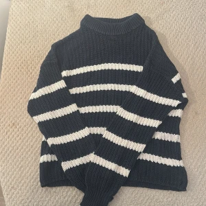 Randig stickad polotröja i mörkblått - Säljer en stickad polotröja i mörkblått med vita horisontella ränder. Tröjan har en hög krage och långa ärmar, perfekt för kyliga dagar. Den grova stickningen ger en mysig och trendig look.