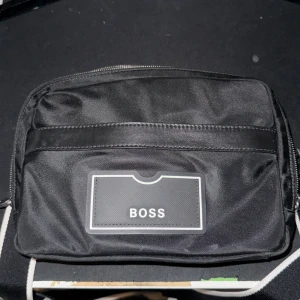 Svart axelväska från BOSS - Snygg svart axelväska från BOSS med justerbar rem och flera fack med dragkedja. Väskan har en stilren design med BOSS-logga framtill och är tillverkad i slitstark polyester. Perfekt för att bära med sig det viktigaste i vardagen.