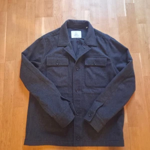 Mörkgrå overshirt Bläck - Stilren mörkgrå overshirt från Bläck med klassisk krage och två bröstfickor med knappar. Jackan är tillverkad i ullblandning och har knäppning framtill samt långa ärmar med knapp vid ärmslut. Perfekt lager-på-lager-plagg för kyligare dagar. Storlek M, säljer då den är för liten nu. Den har inga defekter topp skick. Skriv för frågor och våga skicka pris förslag