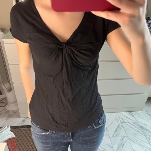 Svart v-ringad topp från Hugo Boss - Svart topp från Hugo Boss med snygg v-ringning och twistad detalj framtill. Modellen har korta ärmar och är figurnära med mjukt, tunt material som känns skönt mot huden. Perfekt för dig som gillar stilrena och enkla plagg med en liten twist.