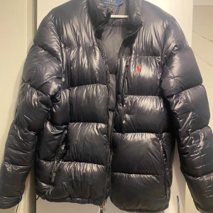 Svart pufferjacka från Polo Ralph Lauren - ! Svart pufferjacka från Polo Ralph Lauren med röd broderad logga på bröstet. Jackan har quiltad design, hög krage och dragkedja framtill samt fickor med dragkedja. Materialet är glansigt och känns riktigt mjukt och skönt. Storlek L men passform mer mot M