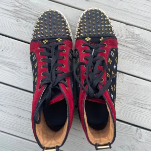 Christian Louboutin sneakers med nitar - Unika high-top sneakers från Christian Louboutin med röd mocka, svarta paneler med broderade guld- och silvermönster samt ikoniska nitar på tån. Skorna har svarta skosnören, vit sula och den klassiska röda undersulan. Lyxig och edgy design som sticker ut. Självklart äkta