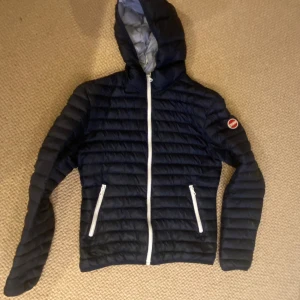 Mörkblå Colmar pufferjacka med huva - Säljer en mörkblå pufferjacka från Colmar Originals med huva och vita dragkedjor. Jackan har ett quiltat mönster och Colmar-logga på ärmen. Perfekt för kyliga dagar och har två praktiska fickor med dragkedja framtill. Några små hål i dunen, men är enkelt att laga och inget som syns om man inte tittar noga. 