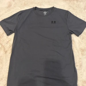 Mörkgrå t-shirt från Under Armour - Snygg mörkgrå t-shirt från Under Armour med klassisk rund hals och svart logga på bröstet och ryggen. Modellen är loose fit och har korta ärmar. Perfekt för dig som gillar sportig och enkel stil.