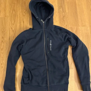 Marinblå hoodie från Sail Racing - Snygg marinblå hoodie från Sail Racing med hel dragkedja och huva med snörning. Sparsamt använd. Pris går att diskutera,