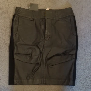 Svart pennkjol från House of Lola - Snygg svart pennkjol från House of Lola med klassisk passform och bälteshällor. Kjolen har fickor fram och bak samt en dekorativ svart sammetsremsa längs sidan. Materialet är mjukt och har en lätt glansig finish, perfekt för en stilren look.