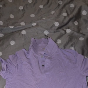 Lila pikétröja från BOSS, storlek M - Säljer en snygg lila pikétröja från BOSS i storlek M. Tröjan har klassisk krage, två knappar framtill och diskret BOSS-brodyr på bröstet. Tillverkad i mjuk bomull som känns skön mot huden. Perfekt för dig som gillar stilrena och trendiga plagg.