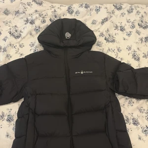 Svart Sail Racing pufferjacka med huva - Svart pufferjacka från Sail Racing med stor huva och logga på bröstet och huvan. Jackan har quiltad design, dragkedja framtill och flera innerfickor. Fylld med dun och har en clean, oversized look som är perfekt för kalla dagar.