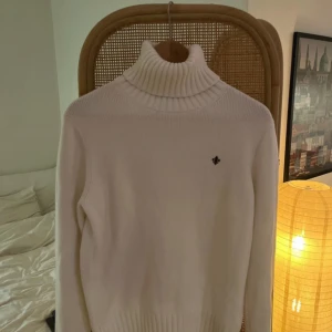 Morris tröja  - Vit polotröja från Morris med ribbad hög krage och diskret broderad logga på bröstet. Tröjan är stickad i mjukt material och har långärmade ärmar. Perfekt för dig som gillar stilrena och klassiska plagg med snygga detaljer.