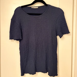 Mörkblå ribbad t-shirt från Zara - Säljer en mörkblå t-shirt från Zara i storlek S. T-shirten har ett ribbat, texturerat tyg och rund halsringning. Modellen är kortärmad och har en loose passform med vågiga kanter vid ärmslut och nederkant. Perfekt för dig som gillar enkel men cool stil.