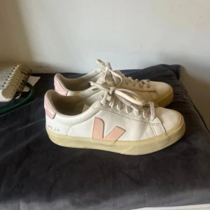 Vita Veja sneakers med rosa detaljer - Snygga vita sneakers från Veja med ljusrosa V-logga på sidan och rosa hälparti. Skorna har klassisk rund tå, snörning och platt sula. Tillverkade i skinn och passar perfekt till en clean och avslappnad stil.