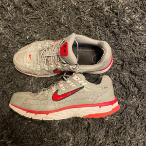 Nike P-6000 grå/röda sneakers - Jag säljer ett par Nike P-6000 som är gråa och röda. Finns tecken på användning t.ex slitage i hälen i ena skon ( se bild 4). Detta har jag dock fixat med en laglapp för ett tag sedan och jag har kunnat använda skon som vanligt!   Kan även skicka med en extra laglapp om den skulle ramla av.   Storlek 44 men kan även användas som ett par 43  Pris kan alltid diskuteras!!