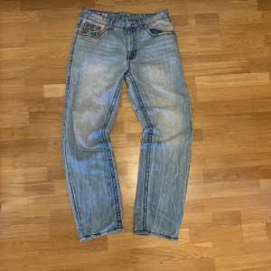 True Religion ljusblå jeans, stl 32 - Säljer ett par ljusblå jeans från True Religion med ikoniska bakfickor och orange kontrastsömmar. Klassisk femficksmodell med dragkedjs gylf och detaljerade logopatchar. Jeansen har en rak passform och är tillverkade i bomull den är använd och har märken på använding som mellan benen som syns på bilden.