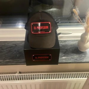 Svart keps John Hatter & Co El Profesor - Svart keps från John Hatter & Co med mesh baktill och justerbar snapback. Framtill sitter en metallskylt med texten 'EL PROFESOR' i rött. Kepsen har en klassisk böjd skärm och cool streetkänsla. Boxen till kepsen är kvar.