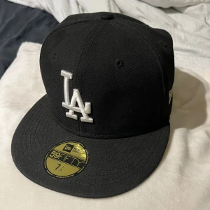 Svart LA-keps New Era 59FIFTY - Snygg svart keps från New Era, modell 59FIFTY, med LA-logga broderad i vitt framtill. Klassisk flat brim och äkta MLB-merch. Kepsen har broderade detaljer på sidan och bak, och är tillverkad i bomull. Den är använd ett fåtal gånger. 