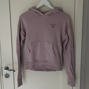 Ljuslila hoodie från GANT - Snygg ljuslila hoodie från GANT som är lite kortare i modellen, med broderad logga på bröstet. Tröjan har huva, magficka och ribbade muddar vid ärmslut och nederkant. Tröjan är använd knappt 5 gånger.
