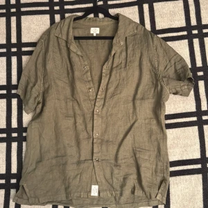 Olivgrön linneskjorta från 1789 Cala - Olivgrön skjorta i linne från 1789 Cala Côte d'Azur, storlek L. Skjortan har korta ärmar, klassisk krage och knappar framtill. Luftig och chill passform, perfekt för varma dagar. Snygg att bära öppen över en t-shirt eller stängd för en clean look.