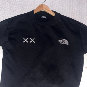Svart sweatshirt från The North Face - Svart sweatshirt från The North Face med broderad logga på bröstet och två vita XX-detaljer. Tröjan har rund hals och långa ärmar, tillverkad i mjuk bomullsmix som är skön att bära. Perfekt för dig som gillar streetwear och enkel stil.