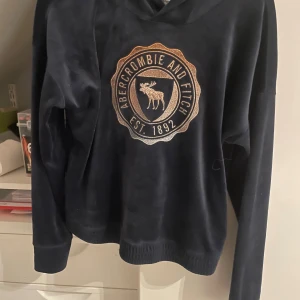 Mörkblå hoodie Abercrombie Kids - Snygg mörkblå hoodie från Abercrombie Kids med stort broderat guldigt emblem och älg på bröstet. Tröjan har huva, långa ärmar och är gjord i mjukt tyg . Den är bra för mig i armarna men lite kort i magen även fast den är 11/12 år (är 170cm) 