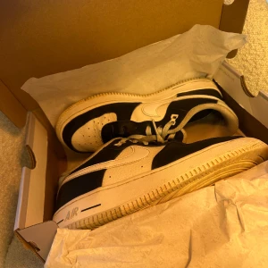 Nike Air Force 1 svart/vit 38.5 - Säljer ett par Nike Air Force 1 '07 i svart och vitt skinn. Klassisk low-top modell med perforerad tå, vit sula och svarta detaljer. Ikonisk Swoosh-logga på sidorna och snörning framtill. Perfekta sneakers för dig som gillar streetstyle.