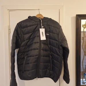Svart pufferjacka från Obey med huva - Svart pufferjacka från Obey med huva och broderad logga på bröstet. Jackan har quiltad design, två sidofickor med dragkedja och hel dragkedja framtill. Perfekt för kalla dagar och har en clean, modern look som funkar till jeans och sneakers.