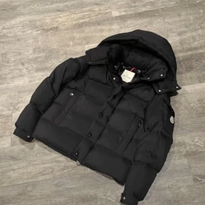 Svart dunjacka från Moncler - Säljer en svart dunjacka från Moncler (vezere) med huva och klassisk logga på ärmen. Jackan är quiltad och har dragkedja samt knappar framtill. Insidan har Monclers ikoniska serietidningsetikett. Perfekt för kalla dagar och riktigt snygg streetstil.