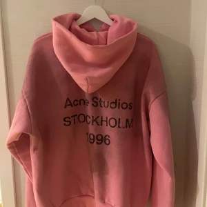 Rosa hoodie från Acne Studios - Säljer en oversized rosa hoodie från Acne Studios med tryck på ryggen: 'Acne Studios STOCKHOLM 1996'. Hoodien har stor huva och är tillverkad i mjuk bomull. Perfekt för dig som gillar streetwear och vill sticka ut med en färgstark look.