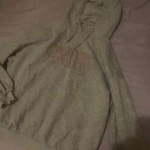 Grå Stranger Things hoodie - Mysig grå hoodie med Stranger Things-tryck i rött på bröstet. Tröjan har huva med snörning och långa ärmar. Perfekt för dig som gillar serien och vill ha en chill och bekväm stil.