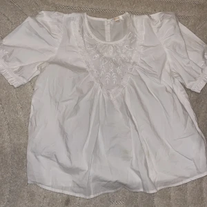 Vit broderad blus från Gina Tricot - Säljer en vit blus från Gina Tricot med korta puffärmar och rund hals. Framsidan har snygga broderade detaljer och hålmönster som ger en romantisk vibe. Blusen är gjord i mjuk bomull och har en knapp i nacken för enkel påklädning.