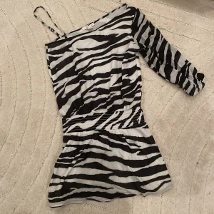 Zebramönstrad kortklänning Gina Tricot - Snygg kortklänning från Gina Tricot x Izaandelle med svartvitt zebramönster. Klänningen har en axelbandslös sida och en lång ärm, vilket ger en cool asymmetrisk look. Perfekt för dig som vill sticka ut med ett djurmönster.