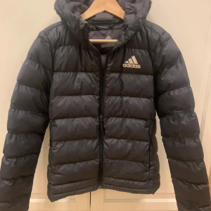 Svart pufferjacka från Adidas XS - Svart pufferjacka från Adidas i storlek XS dam. Jackan har huva, dragkedja framtill och Adidas-logga på bröstet. Den är quiltad och tillverkad i polyester, perfekt för kalla dagar. Snygg och sportig look med normal passform.