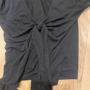 Svart omlottopp från Gina Tricot - Svart långärmad omlottopp från Gina Tricot med knyt framtill och djup v-ringning. Tunn och mjuk i materialet, perfekt att styla med jeans eller kjol. Enkel och stilren design som passar till många tillfällen.