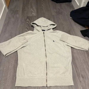 Ljusgrå hoodie från Polo Ralph Lauren - Ljusgrå hoodie från Polo Ralph Lauren med dragkedja och klassisk blå logga på bröstet. Tröjan har huva, kängurufickor och är tillverkad i mjuk bomull. Perfekt för chill dagar eller när du vill ha en clean och enkel look.