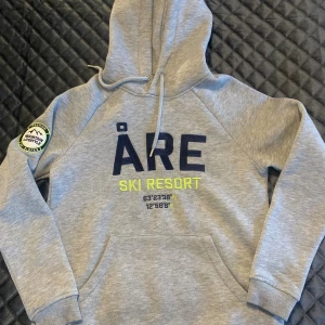 Grå Åre-hoodie från Stadium - Säljer en varm och skön hoodie från Stadium med ÅRE-print framtill. Tröjan har huva med snörning, känguruficka och patch på ärmen. Perfekt för dig som gillar en sportig och chill stil. Är i storlek dam S, men har använts av sonen som stl 146/152. 