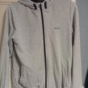 Grå hoodie från Hugo Boss med dragkedja - Snygg ljusgrå hoodie från Hugo Boss med svart dragkedja och huva. Stort BOSS-tryck på ryggen och mindre logga på bröstet. Mjuk bomullsblandning, två fickor fram och långa ärmar. Perfekt för en chill och stilren look. Storlek M men skulle säga den passar också dom s. Pris kan diskuteras 