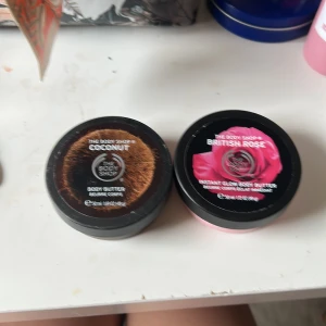 Body butter Coconut & British Rose - Två body butters från The Body Shop, en med kokosdoft i brun burk och en med rosdoft i ljusrosa burk. Båda har krämig, vit konsistens och kommer i runda plastförpackningar med snyggt tryck på locket. Perfekt för dig som älskar mjuk hud och fräscha dofter.