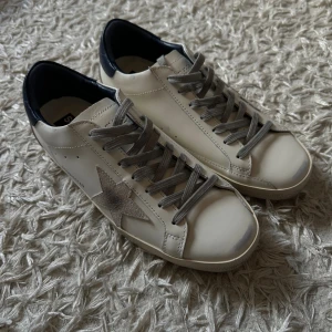 Golden Goose- Sneaker med marinblå tail  - Säljer ett par nya och oanvända Golden Goose  sneakers i skinn med grå mockastjärna på sidan, grå snörning och mörkblå häl. Klassisk rund tå och platt sula. Kommer med all originell förpackning bland annat originalkartongrn de kom i deras dustbag och dess äkthetsbevis. Perfekta för en cool och avslappnad look.  