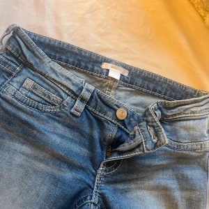ripped bootcut jeans  - Säljer ett par blå jeans från Gina Tricot med slitna detaljer och stora hål på benen. Jeansen har klassisk femficksdesign, dragkedja och knapp framtill samt snygga bakfickor med lock och knapp. Perfekt för dig som gillar en edgy och avslappnad stil. hålen i byxorna är har dock töjts ut så de är ganska stora 