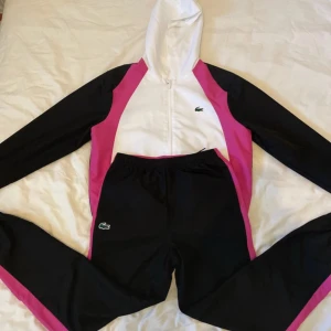 Lacoste tracksuit i svart, vitt och rosa - Säljer en snygg Lacoste tracksuit med dragkedja och huva. Tröjan är svart med vita och rosa paneler, har lång ärm och klassisk Lacoste-logga på bröstet och stor logga på ryggen. Perfekt för dig som gillar sportig och trendig stil.